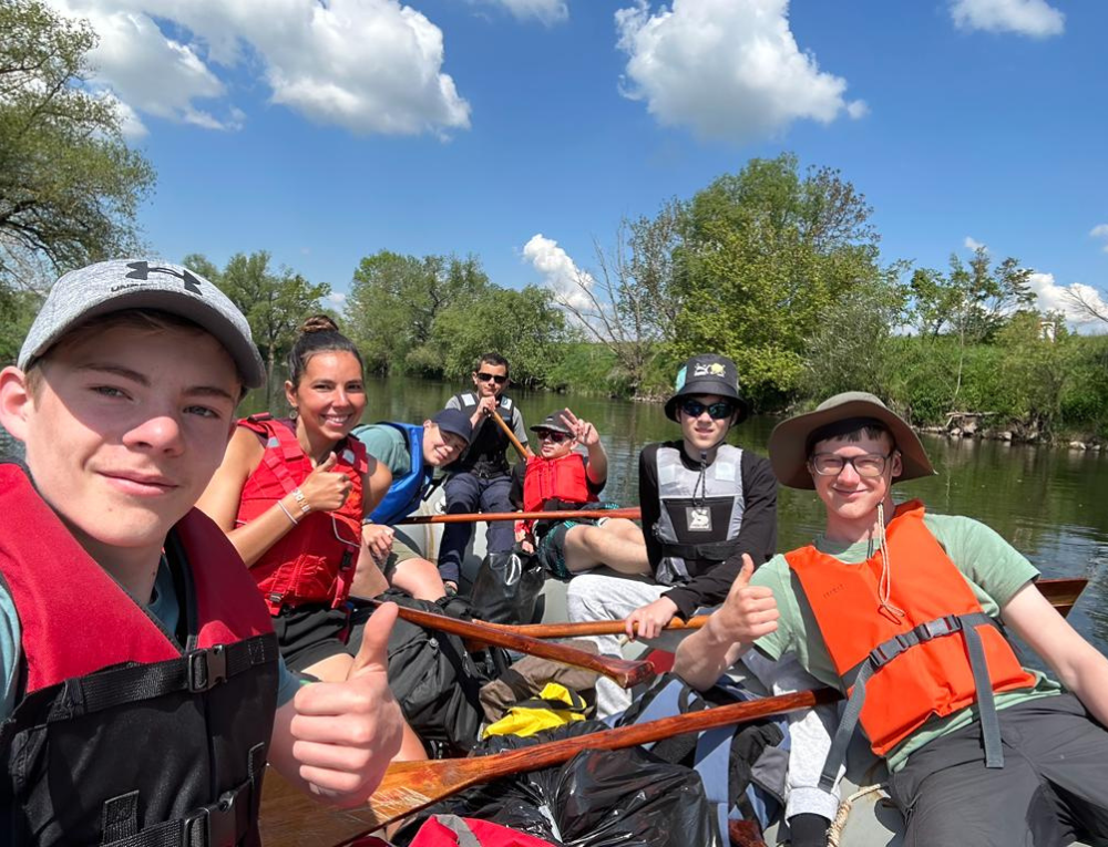 Junge Menschen sitzen freudig zusammen in einem Schlauchboot und lachen in die Kamera. Zudem zeigen einige einen Daumen nach oben.